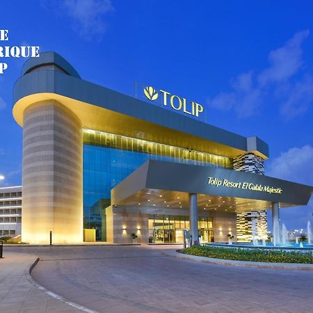Tolip Resort El Galala Majestic Ain Soukhna Exteriér fotografie The photo shows a modern hotel building named "Tolip Resort & Spa," featuring a sleek architectural design with large glass windows. The hotel is illuminated against a twilight sky, showcasing its prominent signage. In the foreground, there is a well