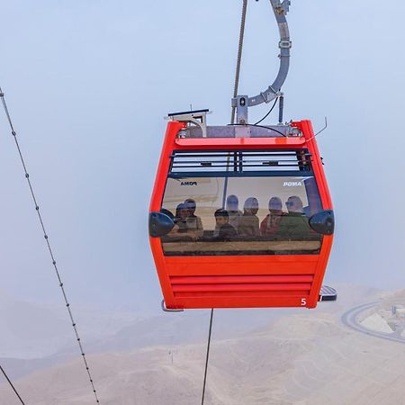 Tolip Resort El Galala Majestic Ain Soukhna Exteriér fotografie The photo shows a red gondola lift suspended in the air, traveling along cables. Inside the gondola, a group of people can be seen. The background features a hazy landscape, possibly mountains or hills, indicating an elevated area. The scene conveys 