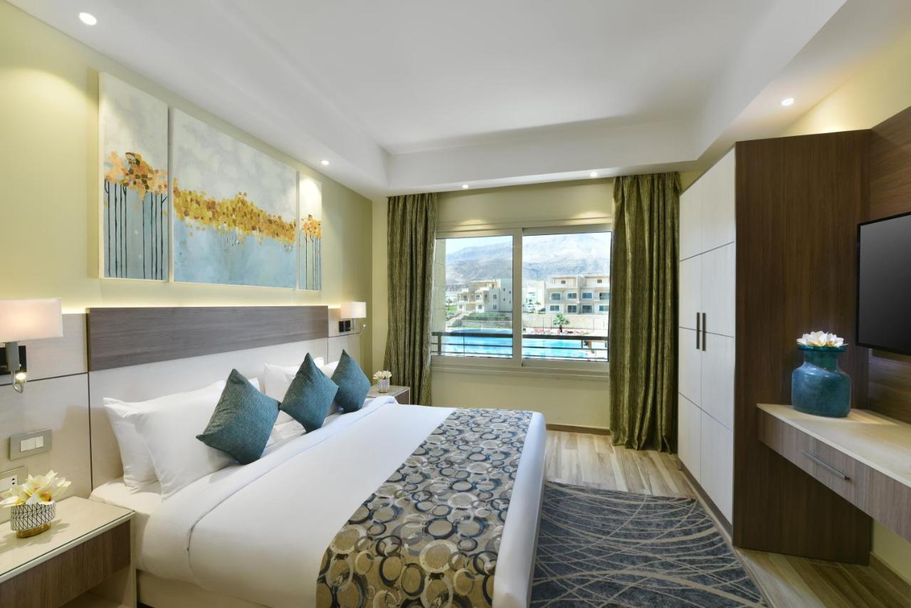 Tolip Resort El Galala Majestic Ain Soukhna Exteriér fotografie The photo shows a modern hotel room. It features a large bed with white bedding and several decorative pillows in shades of teal. Above the bed, there are two pieces of wall art with a landscape theme. The room has a large window with green curtains,