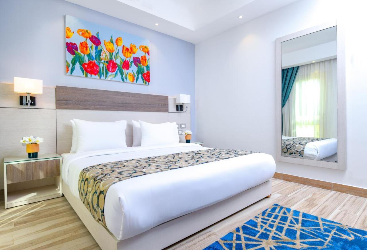 Tolip Resort El Galala Majestic Ain Soukhna Exteriér fotografie The photo shows a modern hotel bedroom. It features a large bed with white linens and a decorative throw blanket. The room has light wooden flooring and is bright, with ample natural light coming from a window. There is a colorful painting of flowers