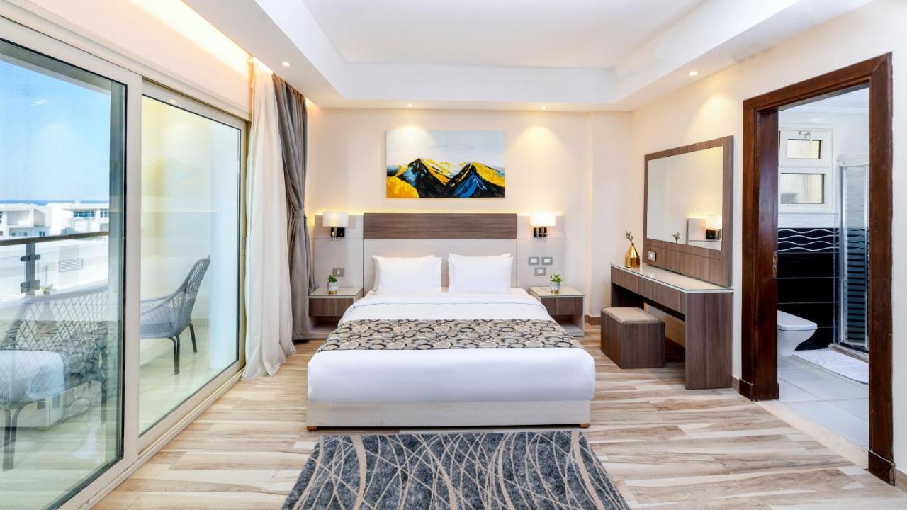 Tolip Resort El Galala Majestic Ain Soukhna Exteriér fotografie The photo shows a modern hotel room featuring a large bed with white linens and a decorative throw. The room has a neutral color palette, accented with warm lighting. There is a piece of artwork above the bed depicting mountains. To the left, there a