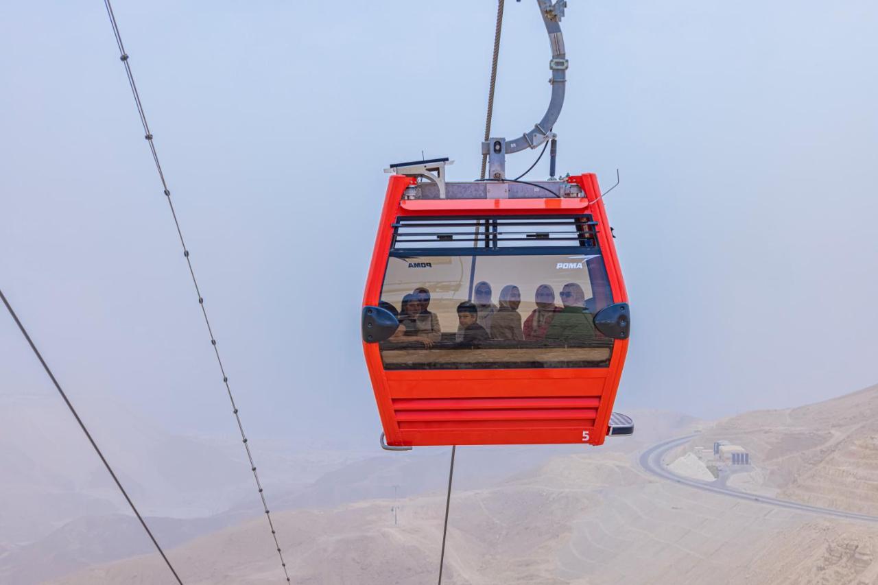 Tolip Resort El Galala Majestic Ain Soukhna Exteriér fotografie The photo shows a red gondola lift suspended in the air, traveling along cables. Inside the gondola, a group of people can be seen. The background features a hazy landscape, possibly mountains or hills, indicating an elevated area. The scene conveys 