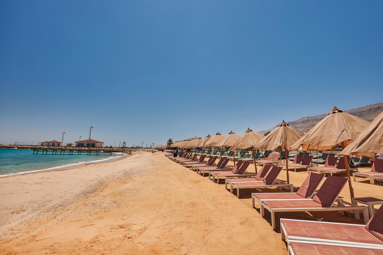 Tolip Resort El Galala Majestic Ain Soukhna Exteriér fotografie The photo shows a scenic beach with a stretch of golden sand under a clear blue sky. There are several beach loungers arranged in rows, each accompanied by thatched umbrellas providing shade. In the background, there is a wooden pier extending into t