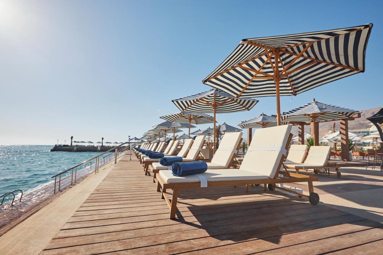 Tolip Resort El Galala Majestic Ain Soukhna Exteriér fotografie The photo depicts a serene beachside setting featuring a row of sun loungers arranged on a wooden deck. Each lounger is equipped with a folded towel and shaded by striped umbrellas. In the background, the water sparkles under bright sunlight, and the