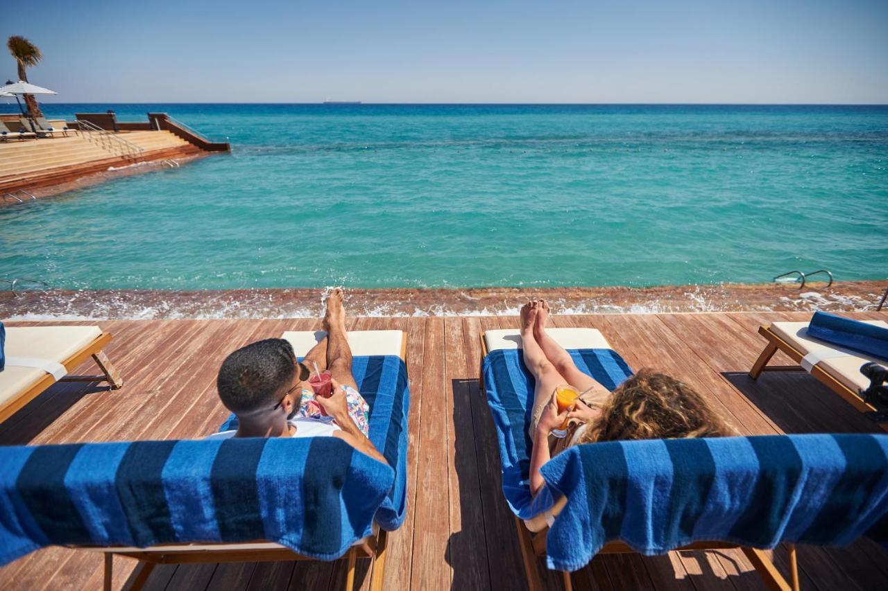 Tolip Resort El Galala Majestic Ain Soukhna Exteriér fotografie The photo depicts two people relaxing on lounge chairs by the waterfront. They are positioned on a wooden deck with a view of the clear, turquoise water. Both individuals are reclining, enjoying the sun, and each has a drink in hand. The scene convey
