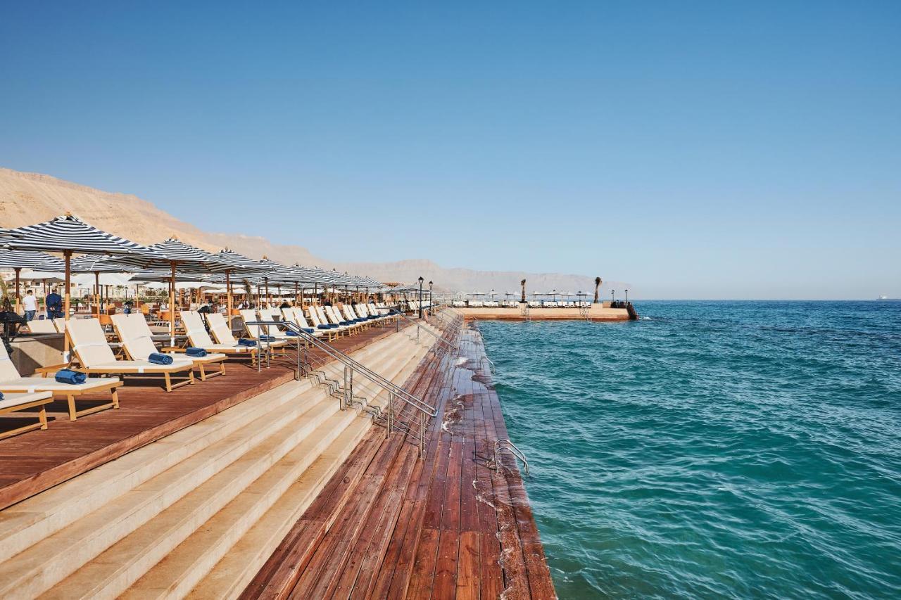 Tolip Resort El Galala Majestic Ain Soukhna Exteriér fotografie The photo shows a seaside area featuring a wooden deck that extends into the water. On the left side, there are several lounge chairs arranged neatly under shade structures, suggesting a relaxing atmosphere. The ocean is visible in the foreground, wi