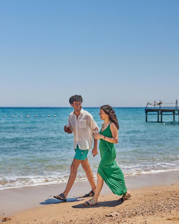 Tolip Resort El Galala Majestic Ain Soukhna Exteriér fotografie The photo shows a couple walking along a beach. The man is wearing a white short-sleeved shirt and green shorts, while the woman is dressed in a flowing green dress. They both appear to be enjoying the pleasant weather, with the ocean in the backgrou