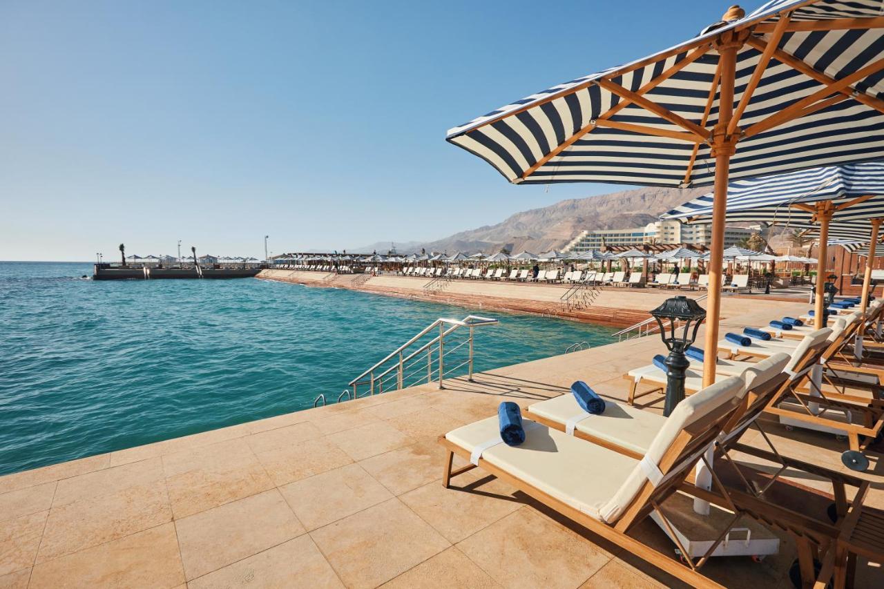 Tolip Resort El Galala Majestic Ain Soukhna Exteriér fotografie The photo shows a scenic beachside area with clear blue water. In the foreground, there are several sun loungers set up on a tiled surface, with blue towels neatly rolled on each one. The loungers are shaded by striped umbrellas. In the distance, a w