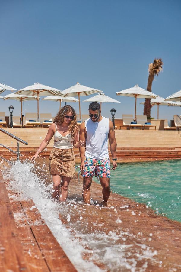 Tolip Resort El Galala Majestic Ain Soukhna Exteriér fotografie The photo depicts a couple walking along a poolside area. They are enjoying a sunny day near a swimming pool, with splashes of water around them. The woman is wearing a light-colored top and a patterned skirt, while the man is dressed in a sleeveless