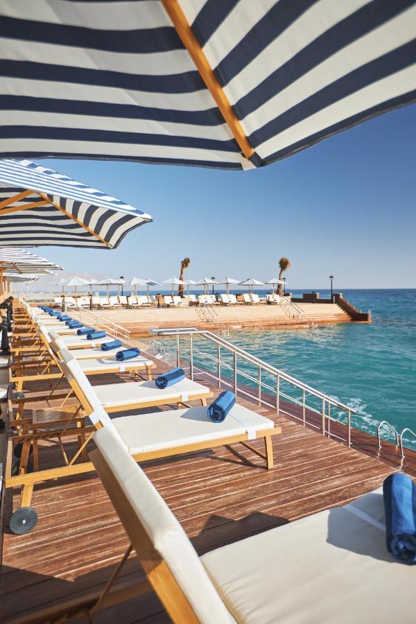 Tolip Resort El Galala Majestic Ain Soukhna Exteriér fotografie The photo depicts a serene outdoor area by the waterfront. There are several lounge chairs arranged in a row, each equipped with blue towels. Large, striped umbrellas provide shade over some of the chairs. In the background, there is a wooden deck th