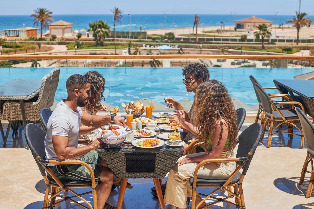 Tolip Resort El Galala Majestic Ain Soukhna Exteriér fotografie The photo shows a group of five people enjoying a meal together outdoors, likely at a restaurant or cafe. They are seated at a table with a beautiful view of a swimming pool and the sea in the background. The table is filled with various dishes, and 