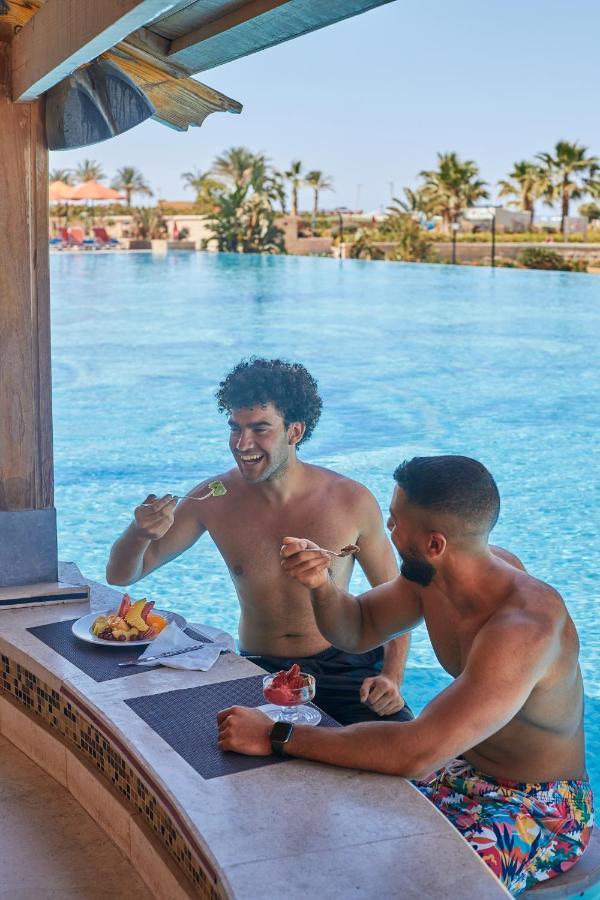 Tolip Resort El Galala Majestic Ain Soukhna Exteriér fotografie The photo features two young men enjoying a meal together. They are sitting at a poolside table with a clear blue swimming pool in the background. The men are shirtless, suggesting a casual, relaxed atmosphere, likely in a warm environment. One man i