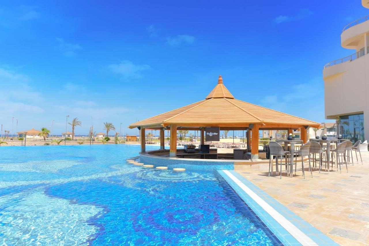 Tolip Resort El Galala Majestic Ain Soukhna Exteriér fotografie The photo shows a beautiful outdoor setting featuring a large swimming pool with clear blue water. In the center, there is a thatched-roof gazebo, which serves as a shaded seating area. Surrounding the gazebo are lounge chairs and a bar area with hig