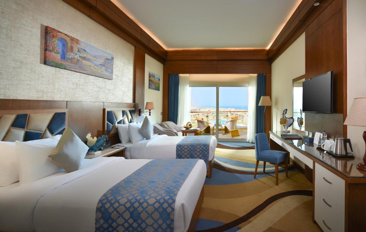 Tolip Resort El Galala Majestic Ain Soukhna Exteriér fotografie The photo shows a well-decorated hotel room featuring two beds with decorative pillows. The room has wooden accents and modern furnishings, including a desk, a television, and lamps. Large windows flood the space with natural light, offering a view o