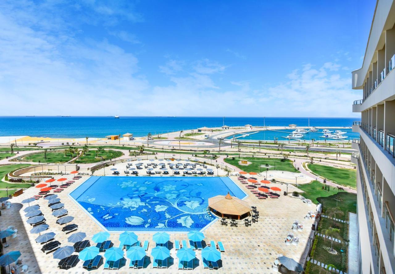 Tolip Resort El Galala Majestic Ain Soukhna Exteriér fotografie The photo shows a beautiful outdoor view of a resort area featuring a large swimming pool with a decorative underwater design. Surrounding the pool are numerous sun loungers and umbrellas in a variety of colors. In the background, there is a scenic c