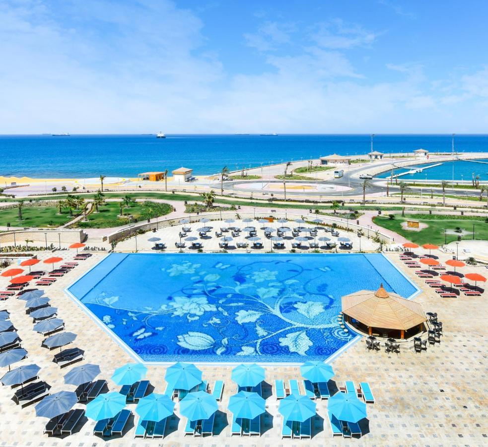 Tolip Resort El Galala Majestic Ain Soukhna Exteriér fotografie The photo features a large swimming pool with a decorative design on its surface, surrounded by sun loungers and umbrellas. In the background, there is a view of the sea, with a clear blue sky and a few scattered clouds. The area is well-maintained, 