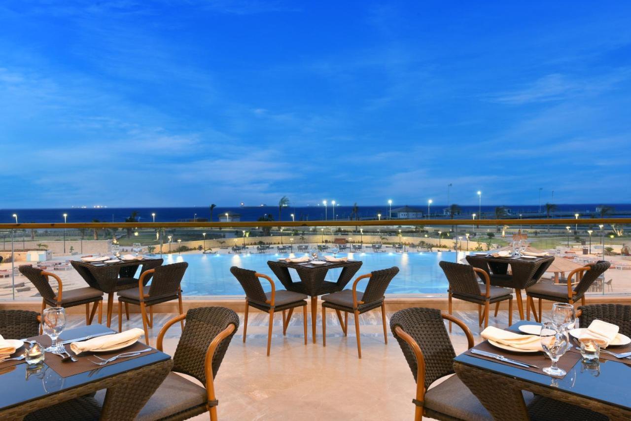 Tolip Resort El Galala Majestic Ain Soukhna Exteriér fotografie The photo shows an outdoor dining area with several neatly arranged tables and chairs. The tables are elegantly set with cutlery and plates, suggesting a fine dining experience. In the background, there is a large swimming pool that reflects the blue