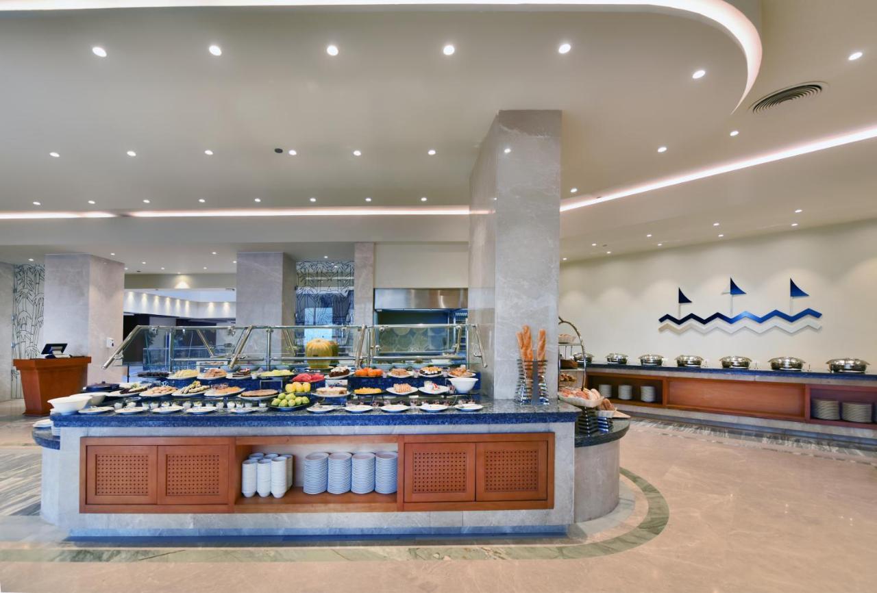 Tolip Resort El Galala Majestic Ain Soukhna Exteriér fotografie The image shows a buffet-style dining area in a restaurant or hotel. There are several food stations with a variety of dishes displayed, including fruits, salads, and other food items. The layout features a large, central island with a wooden base an