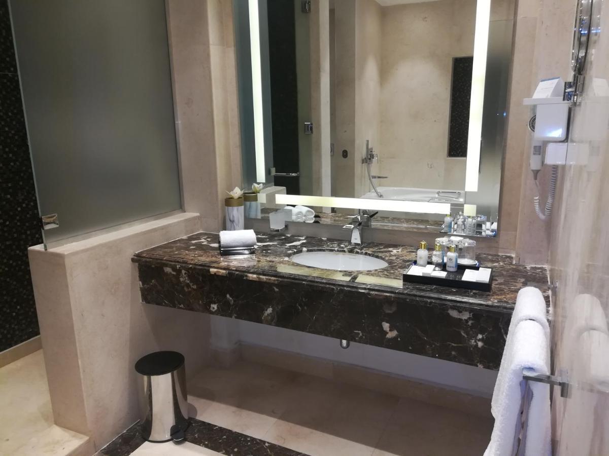 Tolip Resort El Galala Majestic Ain Soukhna Exteriér fotografie The photo shows a modern bathroom with elegant design features. There is a large vanity made of dark marble with a sink and a set of toiletries neatly arranged on top. A mirror with built-in lighting is mounted above the sink. To the side, there is a