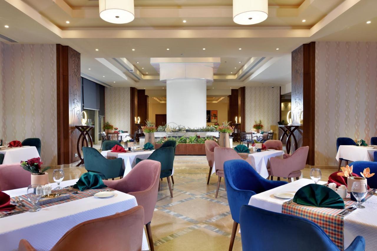 Tolip Resort El Galala Majestic Ain Soukhna Exteriér fotografie The photo shows a spacious and elegantly designed restaurant interior. The layout includes several round tables dressed in white tablecloths, each set with decorative plates, cutlery, and colorful napkins. The chairs are upholstered in a variety of c
