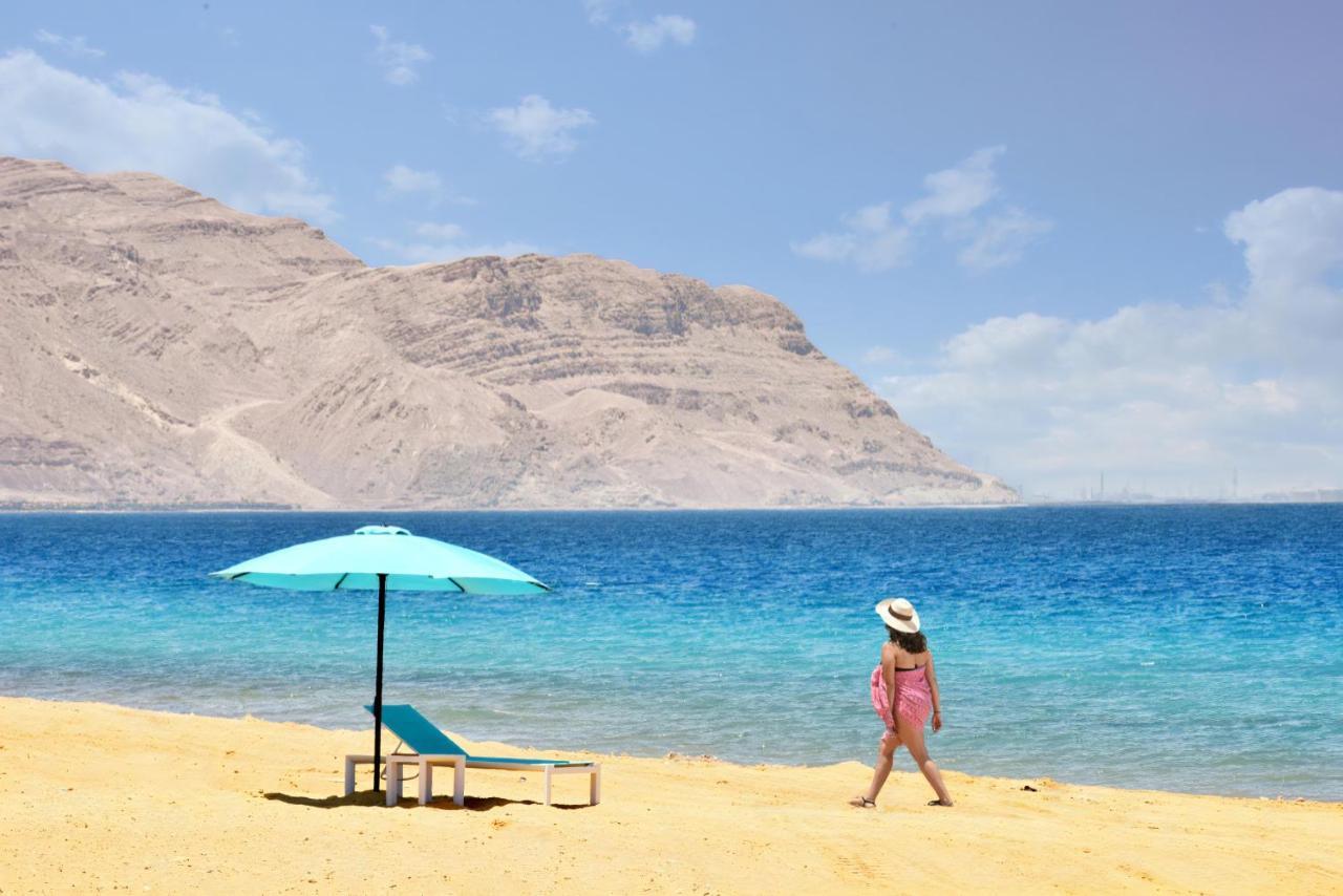 Tolip Resort El Galala Majestic Ain Soukhna Exteriér fotografie The photo depicts a serene beach scene. In the foreground, there is a sandy shore with a single beach chair and an umbrella in a light blue color. A person dressed in a pink outfit is walking along the shoreline, enjoying the beach atmosphere. In the